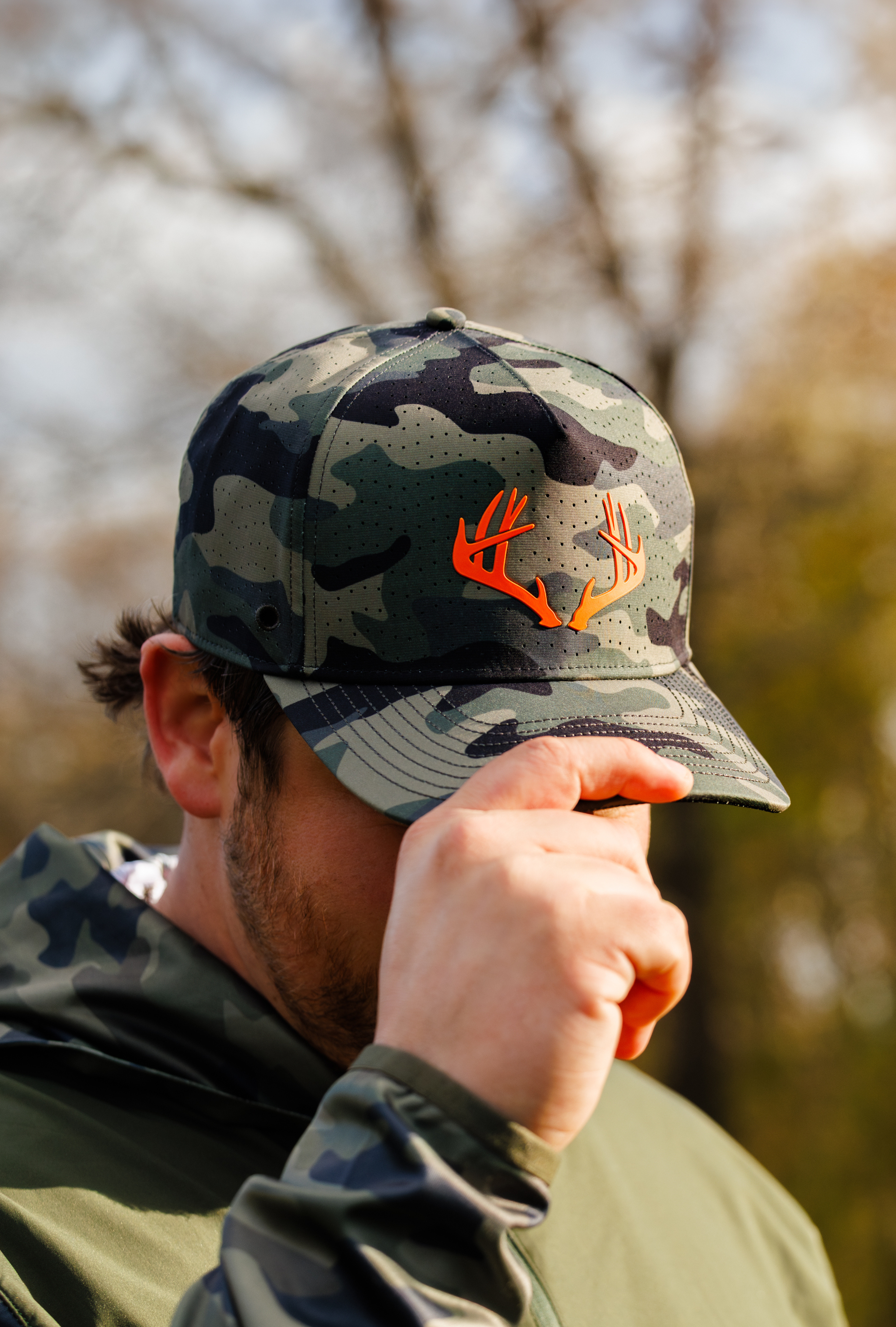 A man outdoors wearing a camo Repel hat with a bright orange antler graphic on the front, paired with a matching camouflage jacket.