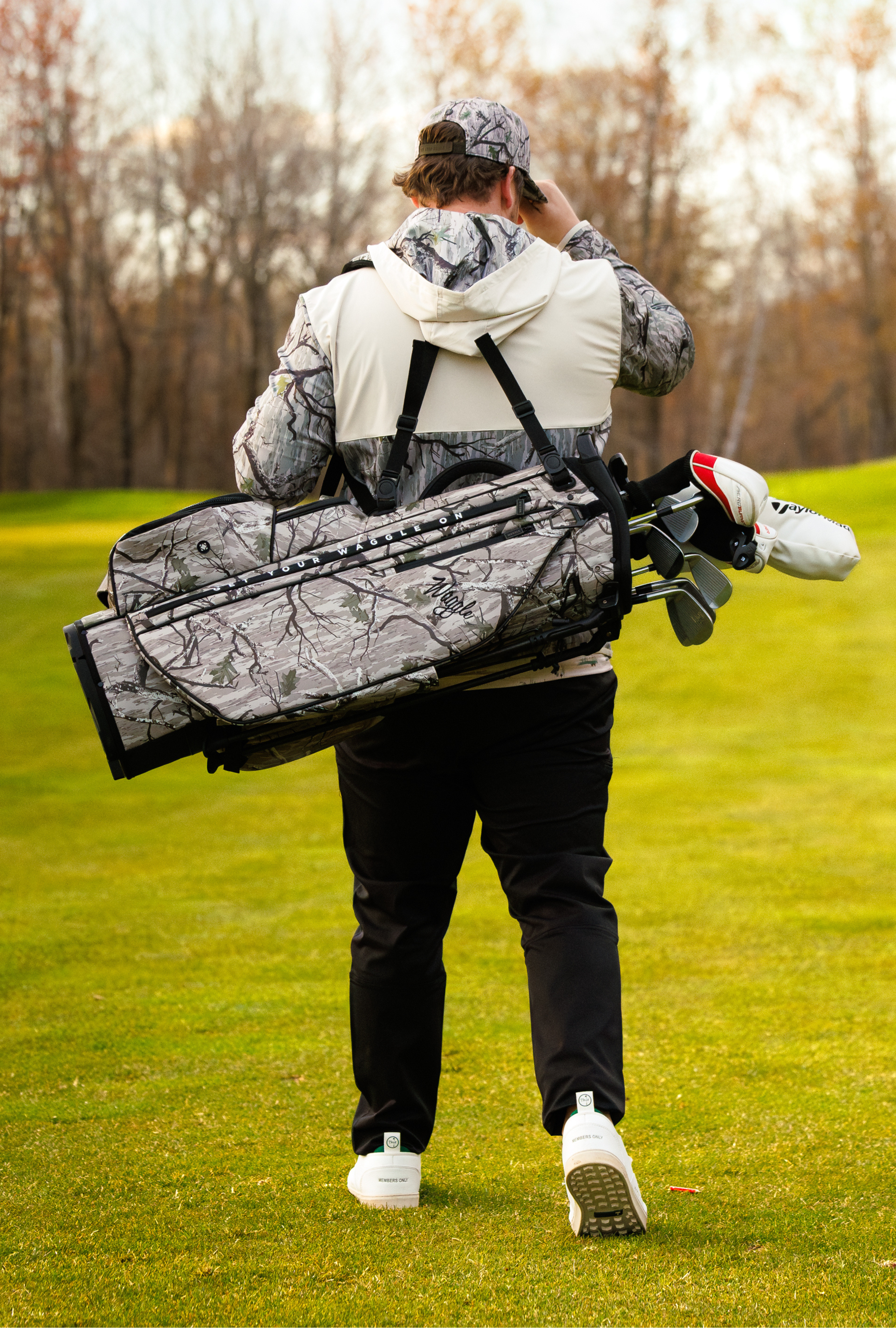 A man on a golf course seen from behind, carrying a camouflage golf bag over his shoulder on a golf course.