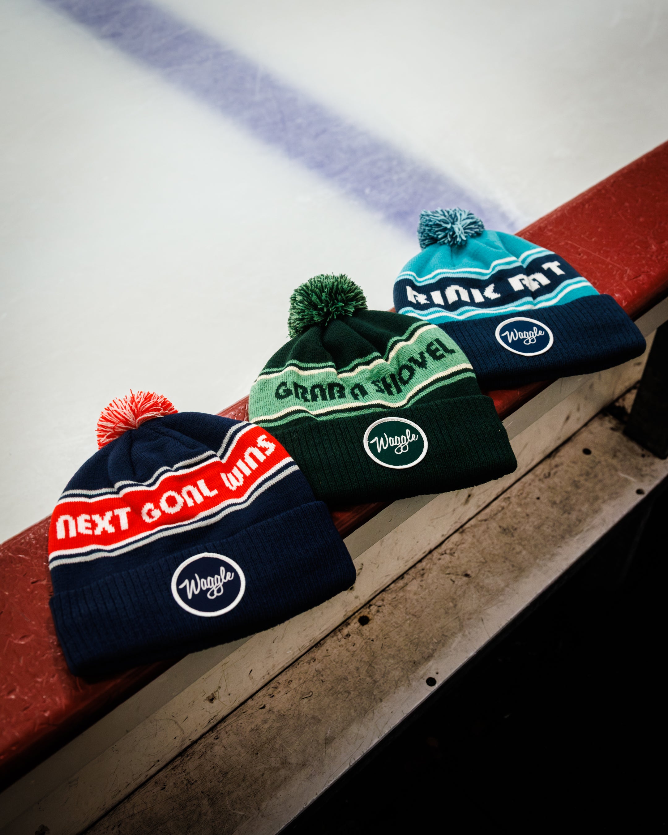 Three pom-pom beanies are lined up on a red boards edge at an ice rink.