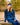 Female model wearing blue crewneck sweater and beanie, sitting on a rock with a nature background.