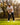 A man wearing a tan polo, black pants, and a hat is holding a golf club on a grassy golf course with trees in the background.