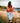 Female Model wearing the Frontier Wildknit Sweater sitting on a rocky shoreline with a lake and trees in the background.