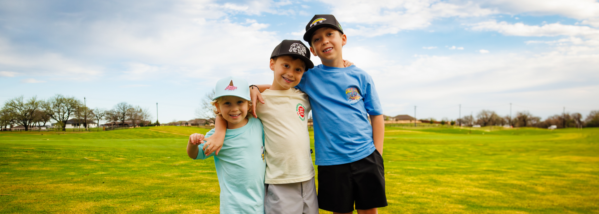 Three children standing together outside, wearing t-shirts and snapbacks.