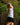 A woman in a golf swing, wearing a white short-sleeve patterned polo and a black skirt, with a blurred outdoor background of green foliage and trees.