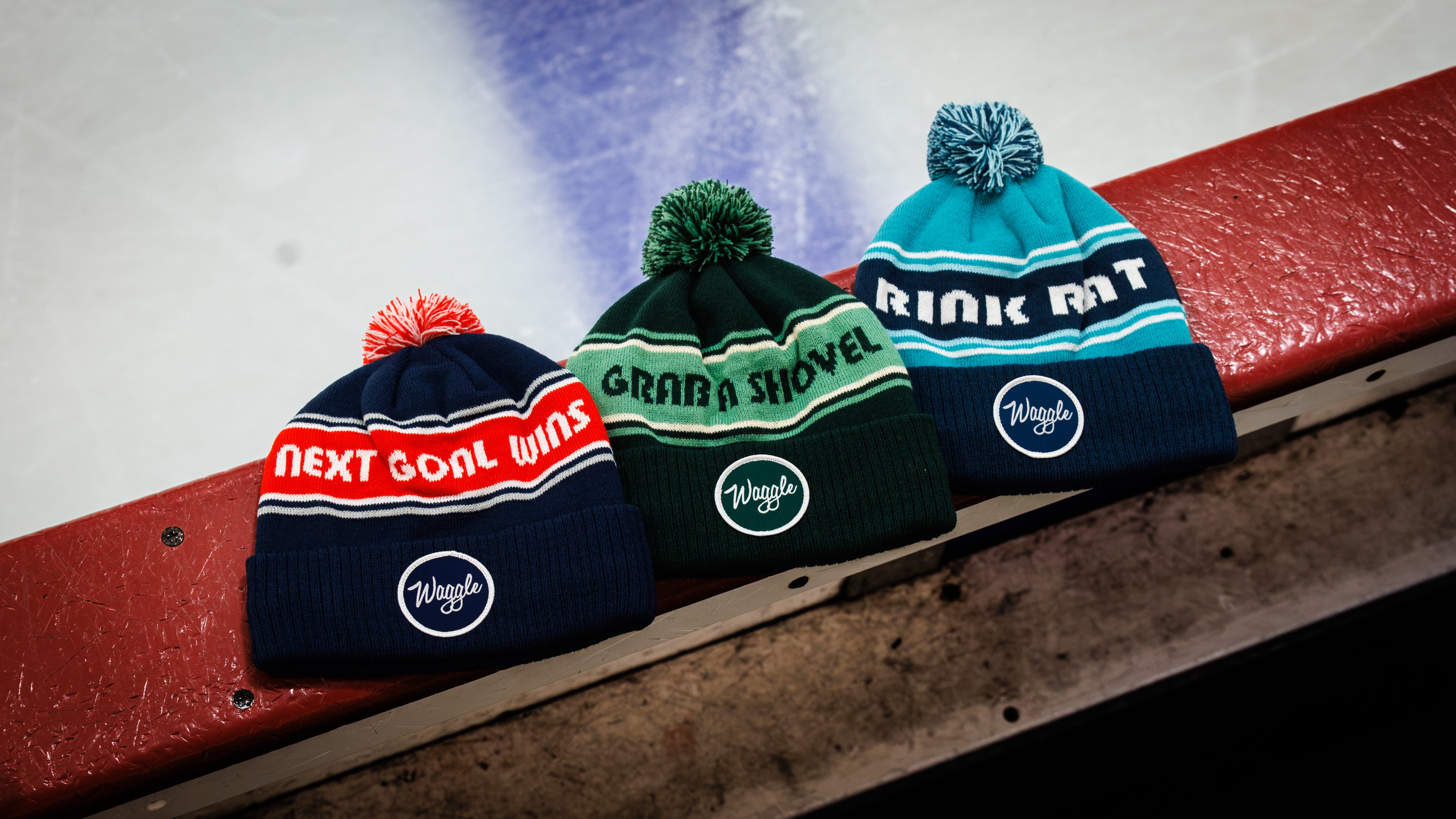 Three pom-pom beanies are lined up on a red boards edge at an ice rink.